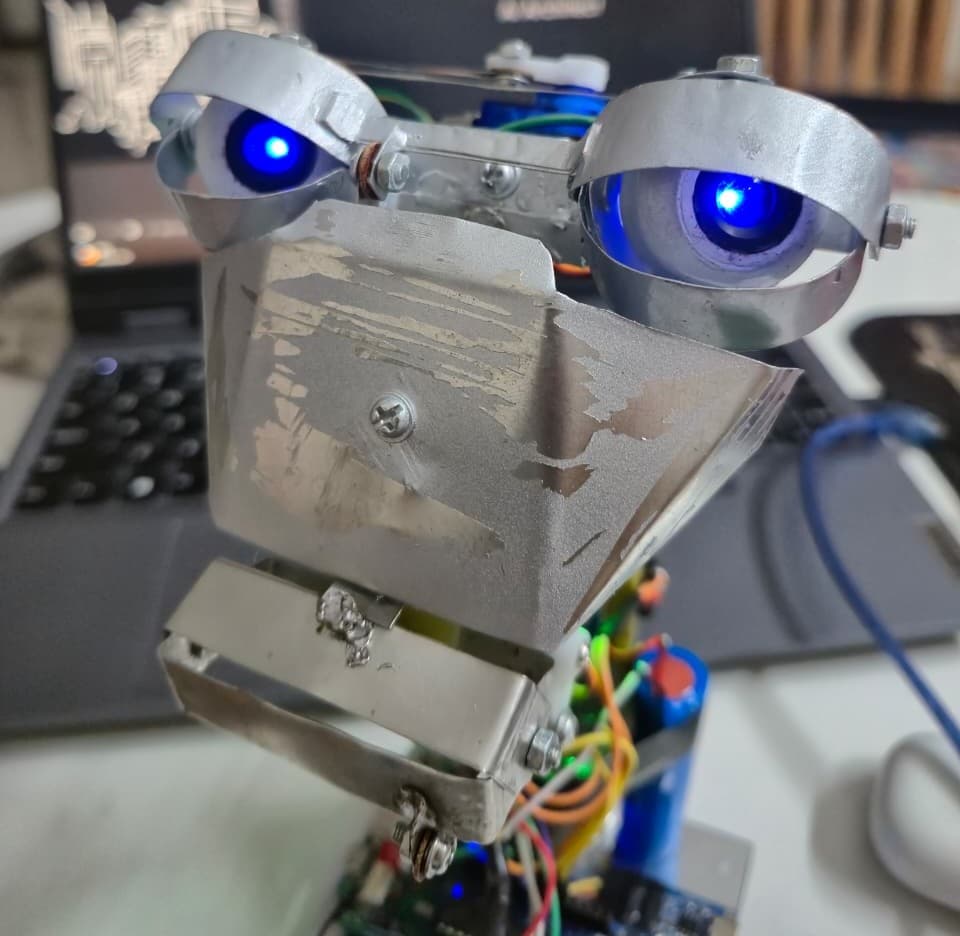 Closeup of the finished robot head: sheet-metal eyelids, glowing blue LED eyes, and the servo wiring and control board exposed below